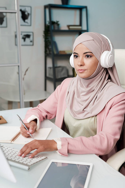 Young serious Muslim woman in headphones, hijab and smart casualwear sitting by table in front of computer monitor and making notes - Foto, Imagen