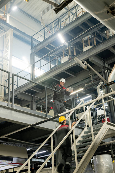 Форман с сетью планшетов внутри большого завода по переработке полимеров, наклоняясь над лестницей, где женщина-работница движется вверх - Фото, изображение