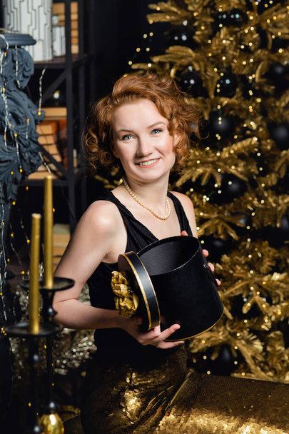 Egy fiatal nő karácsonyi ajándékdobozokkal ajándékokat tart a háttérben egy karácsonyfa arany dekorációval. Stílusos női, divatos estélyi viselet és kiegészítők, smink. Boldog új évet!. - Fotó, kép