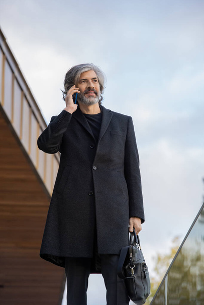 Jistý šedovlasý podnikatel v černém kabátě pomocí smartphonu při konzultaci klienta nebo domluvit schůzku s exteriérem business centra - Fotografie, Obrázek