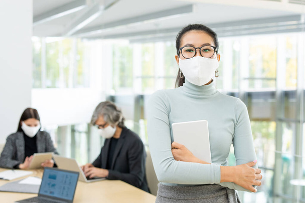Young Asian businesswoman in casualwear and protective mask holding touchpad while standing in front of camera against two colleagues - Photo, Image