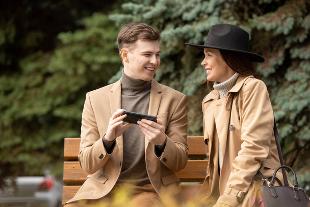 Feliz joven y elegante hombre con teléfono inteligente y su novia sentada en el banco por el árbol de coníferas en el parque y charlando durante el frío - Foto, Imagen
