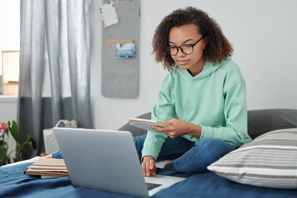 Молодая студентка смешанной расы в повседневной одежде сидит на кровати перед ноутбуком, читает заметки в блокноте и занимается серфингом в сети дома - Фото, изображение