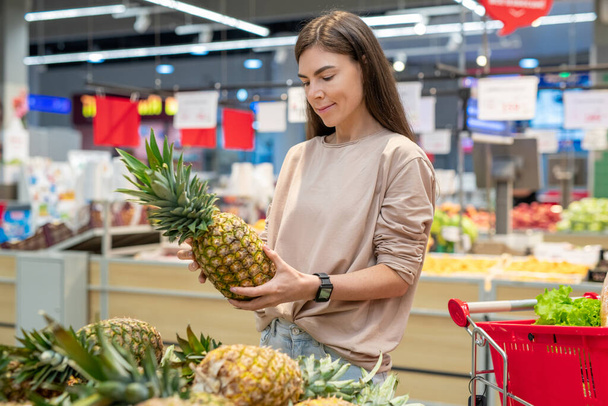 Young woman wearing casual outfit choosing best pinapple in modern supermarket, copy space - Photo, Image