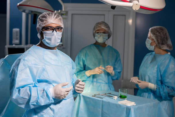 Contemporary mixed-race surgeon in protective mask, gloves and eyeglasses looking at you against his two female assistants - Foto, Imagem