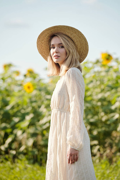 Gorgeous young blond woman in straw hat and white romantic dress standing in front of camera against sunflower field on sunny day - Foto, imagen