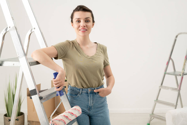 背の高いビューのブルネットの女性でジーンズで座っている上にペイントローラーはしごのステップと見て塗装壁 - 写真・画像