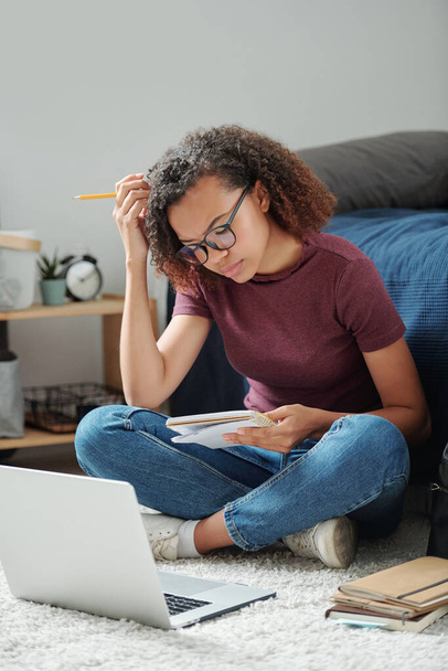 Young serious cross-legged student sitting on the floor by bed in front of laptop and resding her notes in copybook in home environment - Foto, afbeelding