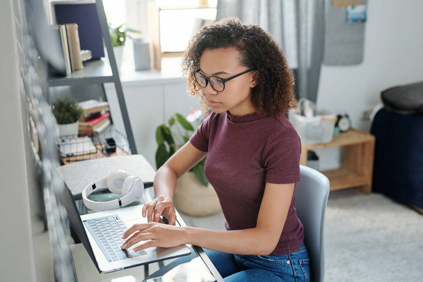 Contemporary young female student in casualwear sitting by small table in front of laptop while taking online course of study in bedroom - Foto, Imagem