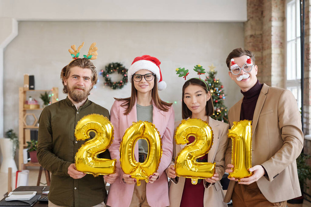 Row of four young intercultural white collar workers in smart casualwear and xmas accessories holding inflatable numbers of next year - 写真・画像