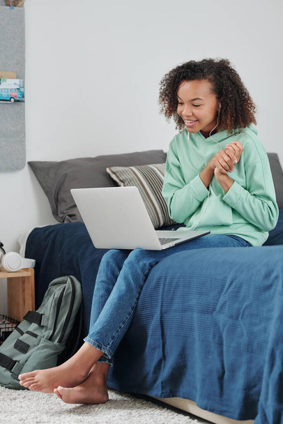 Contemporain gai mixte-race fille dans casulawear et écouteurs avec ordinateur portable sur ses genoux communiquer par chat vidéo à la maison - Photo, image