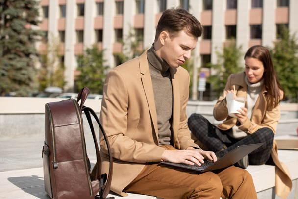Молодой серьезный человек в умной повседневной одежде с помощью ноутбука, сидя на скамейке в городской среде на фоне его девушка ест вок - Фото, изображение