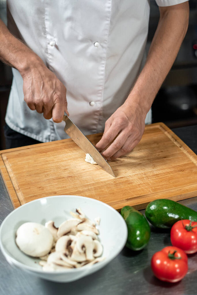 Χέρια του νεαρού αρσενικού σεφ του σύγχρονου εστιατορίου κοπής φρέσκα κρεμμύδια και champignons στο ξύλινο διοικητικό συμβούλιο, ενώ το μαγείρεμα χορτοφαγικό γεύμα - Φωτογραφία, εικόνα