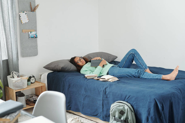 Jeune étudiante épuisée avec livre ouvert sur la poitrine couchée sur le lit et dormant tout en se préparant pour les prochains examens à la maison - Photo, image