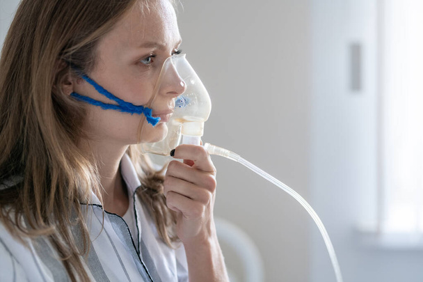 Beteg, fiatal női beteg, aki oxigénmaszkot tart a szája és az orra mellett, miközben levegőt vesz az orvosi rendelőben vagy a kovid kórházban lévő kamrában - Fotó, kép