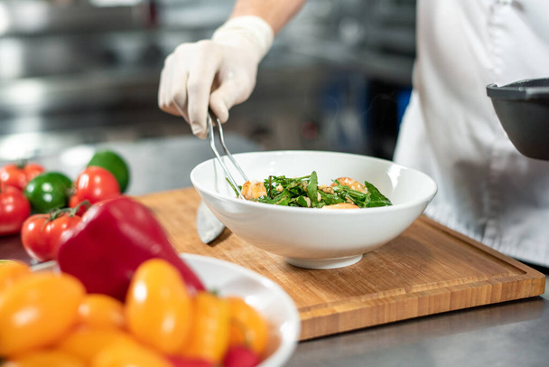 Handbehandschuhte Hand des jungen Kochs mit Gabel, die gekochtes Essen in eine weiße Keramikschüssel legt, die auf einem Holzbrett steht, umgeben von frischem Gemüse - Foto, Bild