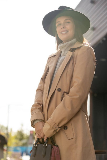 Νέα όμορφη γυναίκα σε μαύρο καπέλο, μπεζ παλτό και γκρι πουλόβερ κοιτάζοντας σας με toothy χαμόγελο, ενώ στέκεται μπροστά από την κάμερα - Φωτογραφία, εικόνα