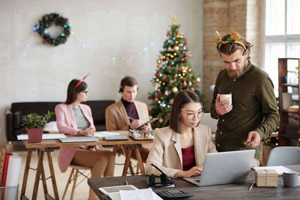 Young confident businessman with xmas headband pointing at laptop display while helping female colleague with preparing presentation - 写真・画像
