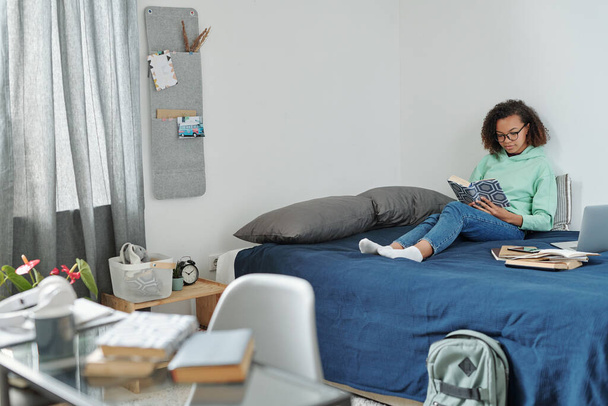 Young pretty diligent student with open book sitting on bed in front of laptop while preparing home assignment in bedroom environment - Photo, image