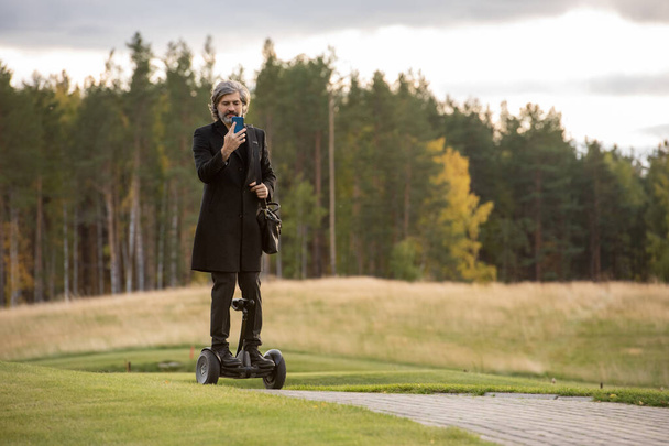 Hombre de negocios contemporáneo de pelo gris con abrigo negro usando un teléfono inteligente mientras está de pie en el giroscopio contra grandes césped verde y árboles - Foto, imagen