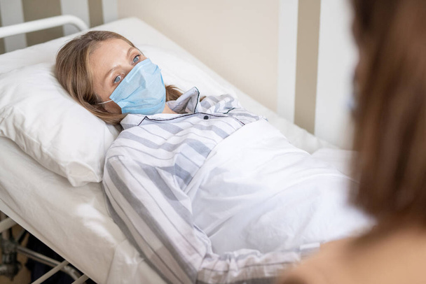 Young blond sick female in pajamas and protective mask lying in bed in hospital chamber and looking at her friend during conversation - Photo, image