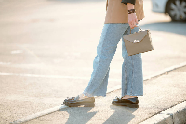 Benen en handen van jonge eigentijdse stijlvolle vrouw in zwarte schoenen, blauwe jeans en bruine jasje die handtas vasthoudt terwijl ze langs de weg staat - Foto, afbeelding