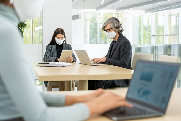 Dwóch zajętych kolegów w inteligentnych ubraniach casualwear i maskach ochronnych siedzących przy stole i korzystających z mobilnych gagdetów podczas pracy w sieci w dużym biurze - Zdjęcie, obraz