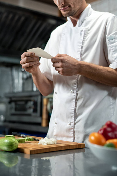 Молодий шеф-кухар в уніформі дивиться через список інгредієнтів для овочевого салату, тримаючи папір над подрібненою цибулею на дерев'яній дошці
 - Фото, зображення