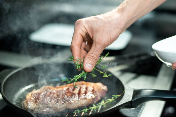 Hand of chef putting green aromatic herbs on roasted piece of meat on hot grill frying pan while standing by electric stove and cooking beef - Foto, imagen