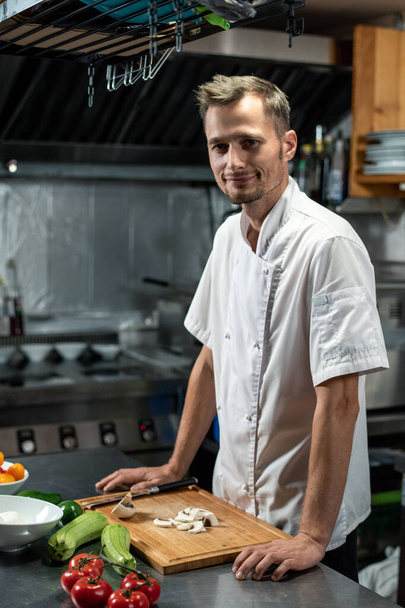 Young successful professional chef of modern restaurant in white uniform standing by table in the kitchen while cooking vegetable coarse - Zdjęcie, obraz