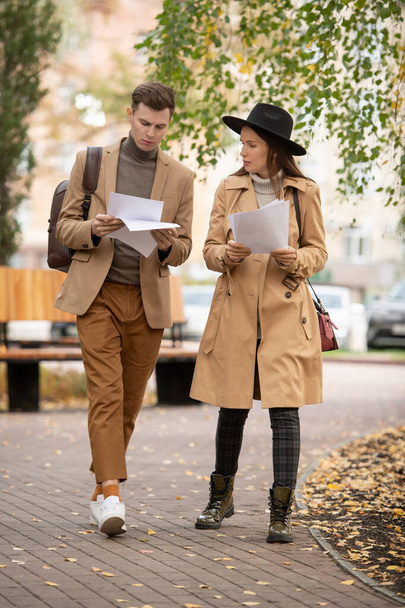 Twee hedendaagse studenten in elegante casualwear lezen kranten en bespreken hun punten tijdens het bewegen langs de weg in het park na de studie - Foto, afbeelding