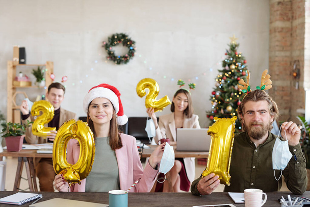 Czterech młodych pracowników biurowych w akcesoriach świątecznych posiadających nadmuchiwane numery w przyszłym roku i maski ochronne, aby pokazać koniec epidemii żywiołowej - Zdjęcie, obraz