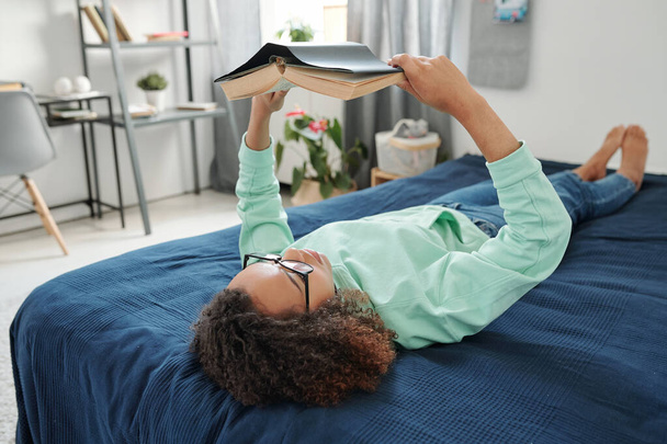 Jonge ontspannen student in een bril en casualwear houden open boek in de voorkant van het gezicht, terwijl liggend op bed en lezen in huis omgeving - Foto, afbeelding