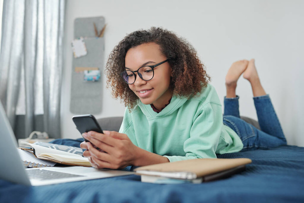 Giovane studentessa sorridente mista in jeans e felpa con cappuccio che scorre nello smartphone mentre è sdraiata sul letto davanti al laptop nella sua camera da letto - Foto, immagini