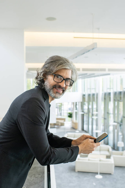 Successful mature businessman in formalwear and eyeglasses looking at you with smile while scrolling in smartphone in business center - Foto, afbeelding