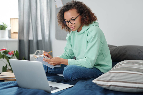 Молода студентка змішаної раси в повсякденному одязі сидить на ліжку перед ноутбуком, роблячи нотатки та слухаючи вчителя під час онлайн-уроку
 - Фото, зображення