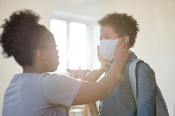 Young careful mother putting protective mask on face of her adorable little son with backpack while seeing him off to school in the morning - Foto, afbeelding