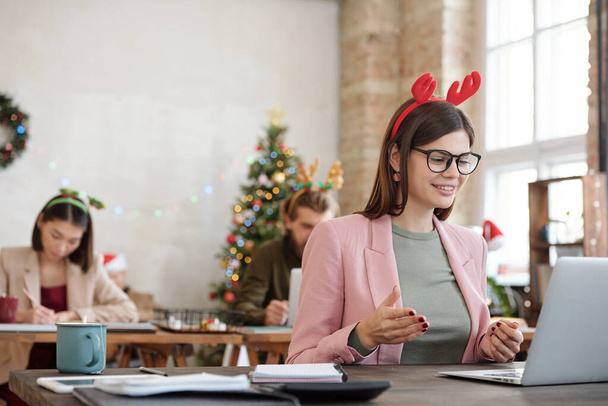 Young cheerful female office manager in eyeglasses, smart casualwear and Christmas headwear talking to client through video chat by table - 写真・画像
