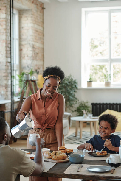 Young happy woman in casualwear pouring coffee or tea into mug of her husband while their cute little son having dessert during breakfast - Photo, Image