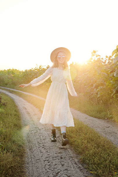 mooi meisje in hoed, zonnebril en jurk bewegen langs land weg tussen zonnebloemen tijdens de promenade in landelijke omgeving op zonnige ochtend - Foto, afbeelding