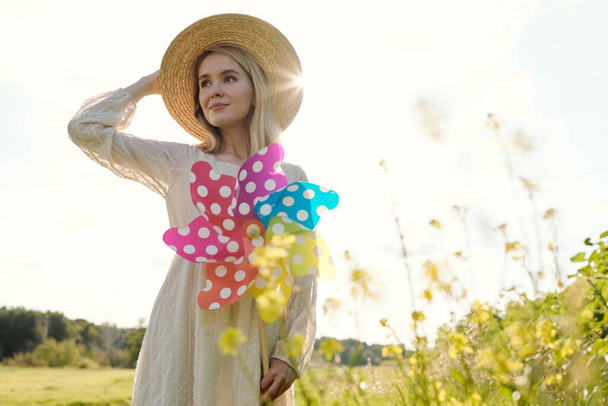 Πανέμορφη νεαρή ξανθιά γυναίκα σε κομψό καπέλο και φόρεμα κρατώντας μεγάλο polkadot whirligig, ενώ στέκεται σε αγροτικό περιβάλλον το καλοκαίρι - Φωτογραφία, εικόνα