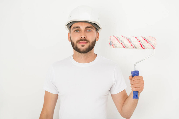 Portrait of smiling handsome young male decorator with stubble wearing hardhat holding paint roll against white wall - Photo, Image