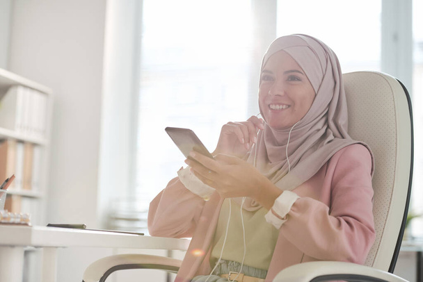 Fiatal mosolygós muszlim üzletasszony hidzsábban és fülhallgató beszél valakivel okostelefonon, miközben ül karosszékben irodai környezetben - Fotó, kép