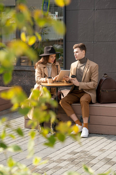 Молодая спокойная пара сидит за маленьким круглым столом по современной архитектуре, пьет и обсуждает новости, а девушка с помощью цифрового планшета - Фото, изображение