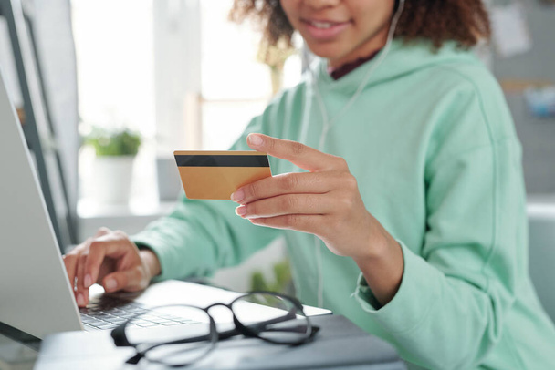 Χέρια της νεαρής γυναίκας μικτής φυλής σε casualwear κάθεται μπροστά από το laptop και πρόκειται να πληρώσει με πιστωτική κάρτα για αγορά στο online κατάστημα - Φωτογραφία, εικόνα