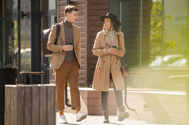 Joven mujer seria y elegante con bebida hablando con su novio en ropa casual caminando cerca en el ambiente urbano durante el frío - Foto, imagen