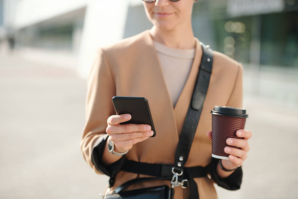 Руки молодої бізнес-леді зі склянкою кави, що прокручується через контакти в смартфоні на відкритому повітрі, ходячи комусь по телефону
 - Фото, зображення