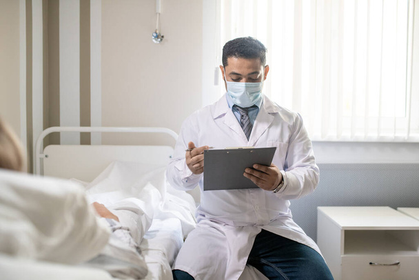 Młody współczesny lekarz w stroju formalnym, białym płaszczu i masce ochronnej czyta dane medyczne w dokumencie, siedząc przy łóżku pacjenta - Zdjęcie, obraz