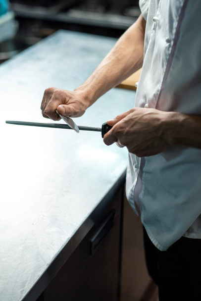 Dłonie młodego współczesnego szefa kuchni restauracji w białym mundurze ostrzenie noża nad stołem kuchennym przed przygotowaniem posiłku - Zdjęcie, obraz