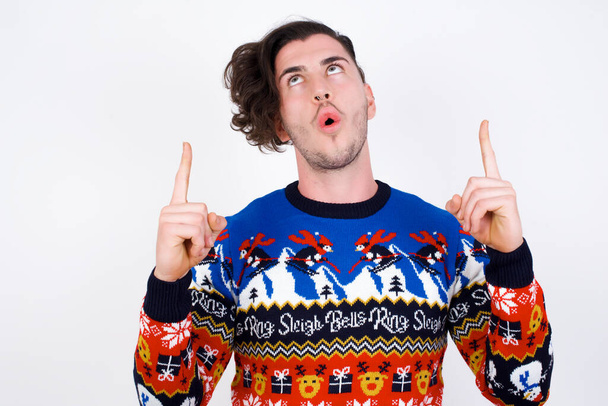  young caucasian man wearing sweater  being amazed and surprised looking and pointing up with fingers showing something strange. - Photo, Image